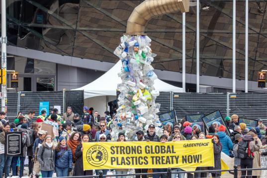 Installation: Aus einem überdimensionierten Wasserhahn tropft Plastik, davor Menschen mit einem Banner "Strong global plastic treaty now".