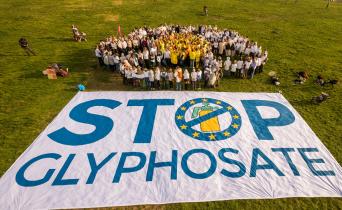Menschen formen eine Blume, im Vordergrund ein großes Banner "Stop Glyphosate"