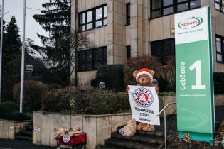 Bärenmarke-Bär mit Nikolausmütze seht vor der Zentrale der Molkerei Hochwald , hält ein Transparent "Mogelmarke. Weidehaltung jetzt!", neben ihm braune Säcke mit Postkarten gefüllt.