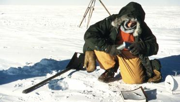 Claude Lorius als junger Mann in der Antarktis. In dicker Kleidung hockt er auf einer endlosen Schneefläche und nimmt eine Probe aus dem Schnee. Neben ihm liegen eine Schaufel, ein Notizbuch und eine Waage.