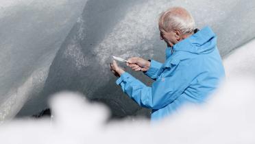 Der 82-jährige Claude Lorius steht in einem Gletscher. In der Hand hält er ein Messer und eine Glasscheibe, auf der er Eis untersucht.