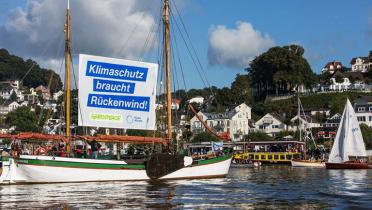 Schiffe fahren auf der Elbe bei Hamburg, eins hat einen Banner mit der Aufschrift "Klimaschutz braucht Rückenwind" gehisst.