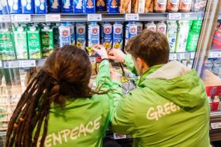 Zei Aktivist:innen in grünen Jacken mit Greenpeace-Schriftzug auf dem Rücken kleben Aufkleber auf Bärenmarke-Milchpackungen