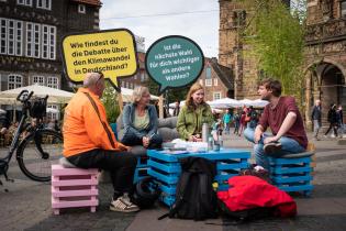 Mann, Frau und zwei Jugendliche in Stadtzentrum auf Hockern vor ausgesägten Sprechblasen mit Fragen für den Zukunfts-Dialog