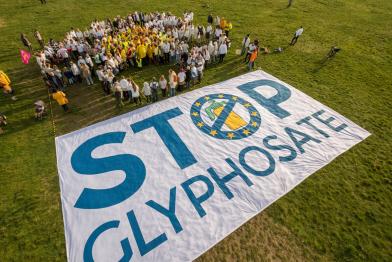 Menschen bilden eine Blume, davor ein großes Banner "Stop Glyphosate"