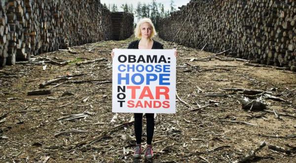 Eine junge Frau steht zwischen zwei riesigen Stapeln aufgeschichteter Baumstämme; sie hält ein Banner mit der Aufschrift "Obama: choose hope not tar sands".
