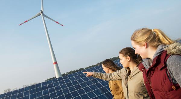 Drei junge Frauen stehen vor einem Solarpanel, dahinter dreht sich ein Windrad. Eine der Frauen zeigt auf die Anlagen.