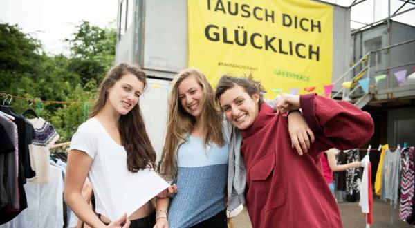 Drei Mädchen präsentieren auf der Kleidertauschparty in Hannover ihre "neuen" Stücke. Über ihnen hängt ein Banner mit der Aufschrift "Tausch dich glücklich"