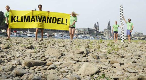 Greenpeace-Aktivisten stehen in der ausgetrockneten Elbe bei Dresden und halten eine große Pegelmesslatte und ein Banner mit der Aufschrift "Klimawandel!" hoch.