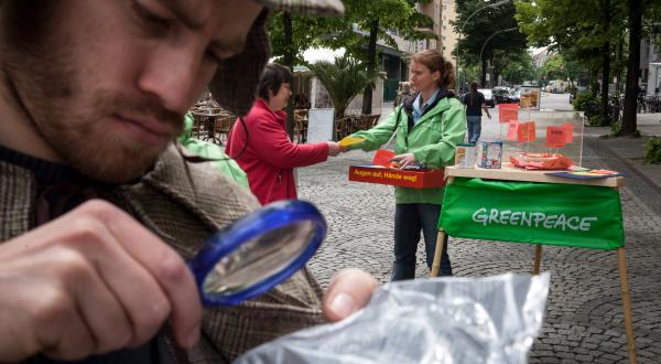 Greenpeace-Aktivisten protestieren im und vor dem KaDeWe (Kaufhaus des Westens) gegen den Verkauf von US-Lebensmitteln, die gentechnisch veränderte Zutaten enthalten. Gen-Detektiv schaut mit einer Lupe auf die kleingedruckte Zutatenliste eines Produkts.