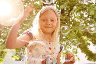 Ein junges Mädchen spielt fröhlich mit Seifenblasen bei schönem Wetter, im Hintergrund sind Bäume zu sehen.
