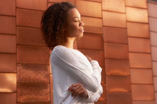 Eine junge Frau steht vor einer rot-braunen Wand im Sonnenlicht und atmet besonnen ein.