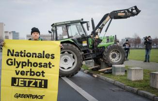 Aktivist hält ein Banner "NAtionales Glyphosatverbot jetzt", im Hintergrund ein Trecker 
