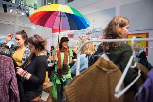 Eine Frau hält einen Regenschirm in Regenbogenfarben, um sie herum Menschen und Kleiderständer