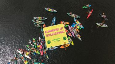 Kajaks, Kanus und Schwimmer:innen im Wasser, auf einem schwimmenden Banner steht: Klimakrise ist Fluchtgrund!