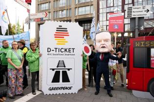 Aktive halten ein überdimensioniertes Deutschlandticket, daneben steht ein:e Aktivist:in mit einer Wissing-Maske