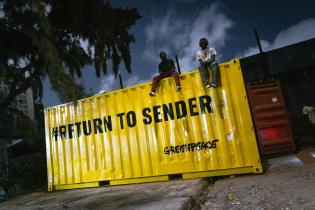 Großer gelber Container mit der Aufschrift "Return to Sender", auf dem zwei Aktive sitzen.