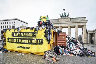 Aktive sitzen auf einem hohen Textilmüll-Berg  vor dem Brandenburger Tor, davor ein Container, auf dem Banner steht "Fast Fashion: Kleider machen Müll".