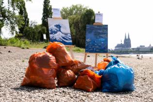 Mit Verpackungsmüll gefüllte Säcke vor zwei Gemälden am Flussufer