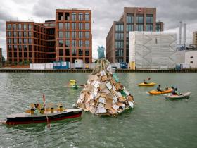 Ein als Titanic verkleidetes Kajak steuert im Hafenbecken auf einen etwa drei Meter hohen Eisberg aus Verpackungsmüll zu. Im Hintergrund Aktive auf Stand-up Paddleboards mit Bannern. 