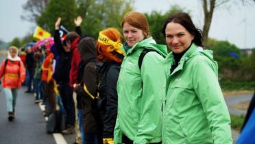 Zwei junge Frauen in grünen Greenpeace-Jacken stehen nebeneinander. Im Hintergrund sieht man weitere Menschen, sie halten sich zum Teil an den Händen. 