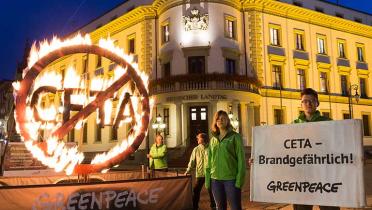 Greenpeace-Aktivisten stehen mit einem drei Meter großen, glühenden CETA-Symbol vor dem Landtag in Wiebaden. „CETA -Brandgefährlich!“ warnt ein Banner neben dem leuchtenden Zeichen.