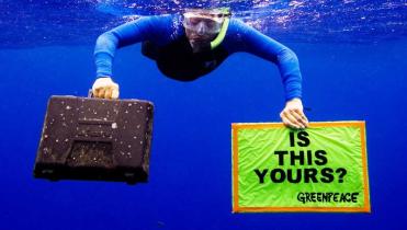 Ein Taucher hält unter Wasser eine Plasik-Box, die er aus dem Meer gefischt hat, und ein Banner mit der Aufschrift "Is this yours?"