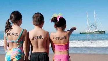 Kinder mit Detox-Schriftzug auf dem Rücken beobachten von einem Strand aus die Rainbow Warrior