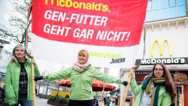Drei Aktivistinnen der Greenpeace-Jugend stehen vor einer McDonald's-Filiale. Sie halten ein rotes Transparent mit der Aufschrift: "Gen-Futter geht gar nicht"
