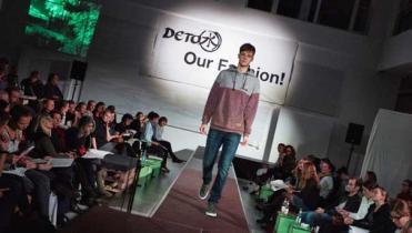 Ein junger Mann führt auf dem Laufsteg in der Hamburger Greenpeace-Zentrale Öko-Mode vor; durmherum sitzen Zuschauer. Der Mann trägt ein grau-rotes Sweatshirt und Jeans; hinter ihm hängt ein weißes Banner mit der Aufschrift "Detox our fashion"