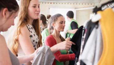Drei junge Frauen stehen vor einer Kleiderstange, auf der jede Menge Klamotten hängen, und begutachten die Kleidungsstücke.