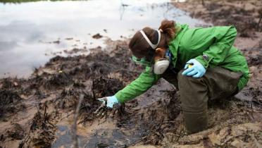 eine Greenpeace-Mitarbeiterin mit Atemmaske hockt vor einer einer Ölpfütze. Sie hat die Finger hineingetaucht; an ihrem weißen Handschuh klebt Öl.