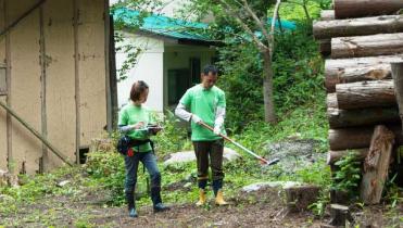 Zwei Greenpeace-Mitarbeiter messen die Strahlung zwischen Häusern in Iitate. Einer hält ein Messgerät an einer Stange; neben ihm liegen aufgeschichtete Baumstämme.