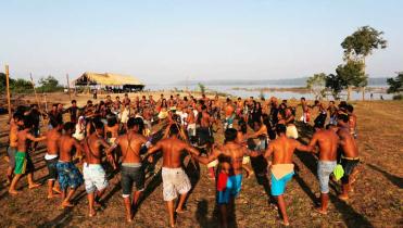 Auf einem Sandplatz am Ufer des Tapajós tanzen Männer des Munduruk-Volkes einen Kreistanz. Sie haben einander mit den Armen untergehakt, tragen Shorts und haben nackte Oberkörper. Im Hintergrund: der Fluss, Bäume, eine offene Hütte mit Palmwedel-Dach.