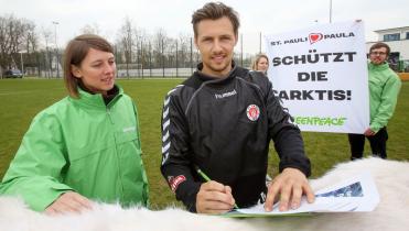 Auf dem Rücken von Eisbär-Dame Paula unterzeichnet Sören Gonther, Mannschaftskapitän des FC St. Pauli, eine Petition zum Schutz der Arktis. Hinter ihm halten Greenpeace-Aktivisten ein Banner mit der Aufschrift "Schützt die Arktis".