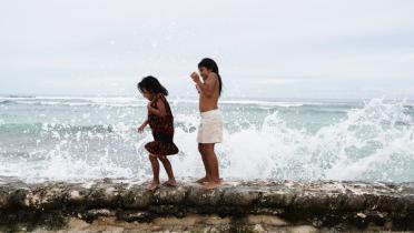 Zwei Mädchen laufen auf einer Mauer entlang und lachen. Die Wellen des Meeres spritzen über die Mauer.