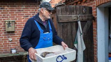 Kleinfischer Wolfgang Albrecht vor seinem Haus, er trägt eine Kiste mit einem Fisch.