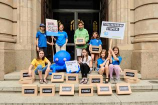 Greenteam-Kinder versammeln sich mit Protestschildern, blauer Krake und Kartons voller Unterschriften gegen den Tiefseebergbau auf den Stufen zum Bundeswirtschaftsministerium.