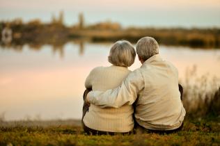 Ein älteres, sich im Arm haltendes Paar sitzt mit dem Rücken gewandt bei Sonnenuntergang am See und blickt auf das Wasser.