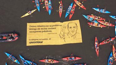 Ein großes schwimmendes Banner zeigt Papst Franziskus mit einem Zitat, umringt von zahlreichen Kajaks