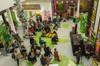 Blick von oben: Viele Menschen sitzen oder stehen um ein Podium in der Greenpeace-Ausstellung und hören einer Frau zu.