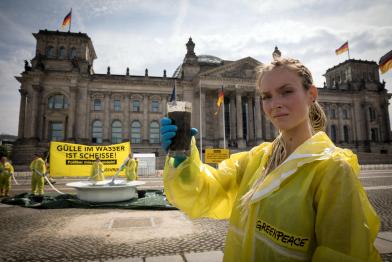 Drei Meter großen Teller voller Gülle vor dem Reichstag in Berlin: Eine Aktivistin hält einen Becher mit dunkler Flüssigkeit. Auf dem Banner steht: „Gülle im Wasser ist Scheiße! Politiker müssen handeln.“
