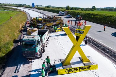 Aktive protestieren an einem zwölf Meter breiten Betonfertiger mit Bannern „Beton stoppen – Natur schützen“ und einem großen gelben X 