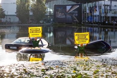 Wasserbecken vor der Messe mit drei eingesunkenen Autodächern. Aktive im Wasser halten Banner: „Autoindustrie versenkt Klimaschutz“ und „Shrink Now Or Sink Later“