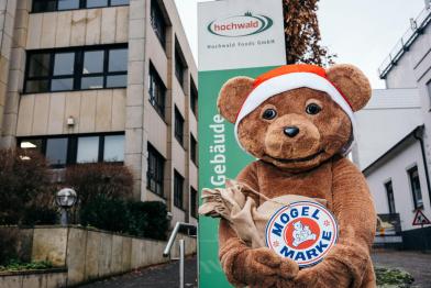 Bärenmarke-Bär mit Nikolausmütze steht vor Firmenzentrale und hält einen braunen Sack.