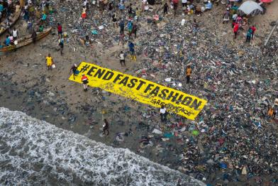 Großes Banner "End Fast Fashion" liegt am Strand in Ghana zwischen Textilmüll.