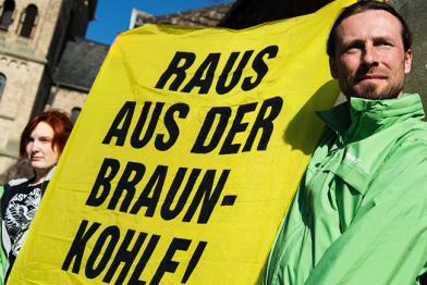 Ein Mann in grüner Greenpeace-Jacke hält gemeinsam mit einer Frau ein gelbes Plakat hoch, auf dem steht: " Raus aus der Braunkohle!"
