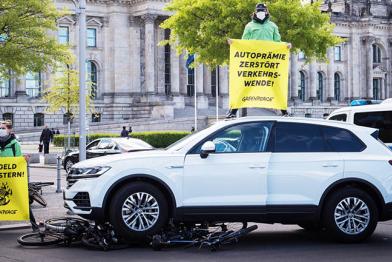 Aktivist*innen stehen auf dem Autodach und halten ein Banner "Autoprämie zerstört Verkehrswende", vor dem Auto liegen überfahrene bereits vor der Aktion schrottreife Räder.