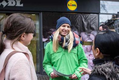 Judith Blümcke (Mitte) von Greenpeace Köln im Gespräch mit Passanten. Im Hintergrund überdecken andere die Fensterscheiben einer Lidl-Filiale von außen mit Fotos aus Schweineställen.