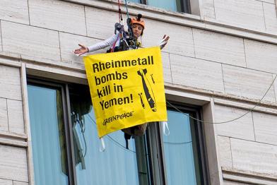 Greenpeace-Kletterin an Hauswand hält Banner mit "Rheinmetalbombs kill in Jemen"